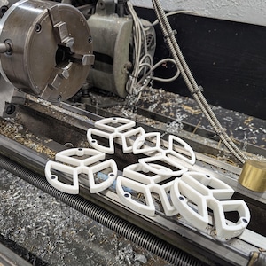 May include: White, geometric, 3D-printed objects, possibly components or parts, are arranged on a metal lathe. The objects have a unique, open design with rounded edges and a three-part structure. A brass cylinder is also visible.