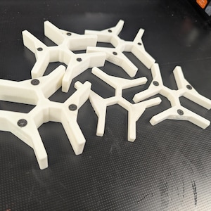 Op de afbeelding: Een verzameling witte, Y-vormige plastic componenten met zwarte schroeven. De componenten zijn gerangschikt in een cluster op een donker, getextureerd oppervlak. Het ontwerp lijkt modulair of in elkaar te passen.