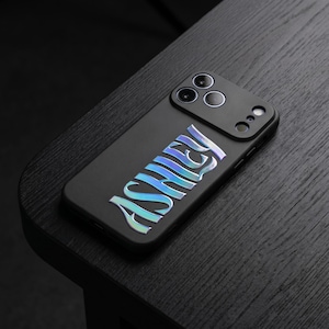 May include: Black phone case with the name "Ashley" in iridescent, holographic lettering. The case has a matte finish and features a camera module with three lenses. The phone case is resting on a dark wooden surface.