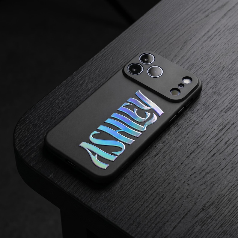 May include: Black phone case with the name "Ashley" in iridescent, holographic lettering. The case has a matte finish and features a camera module with three lenses. The phone case is resting on a dark wooden surface.