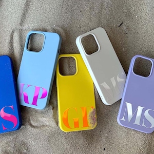 May include: A collection of colorful phone cases in blue, yellow, white, and purple, each with a unique initial design. The cases are arranged on a sandy surface, showcasing a variety of personalized accessories.