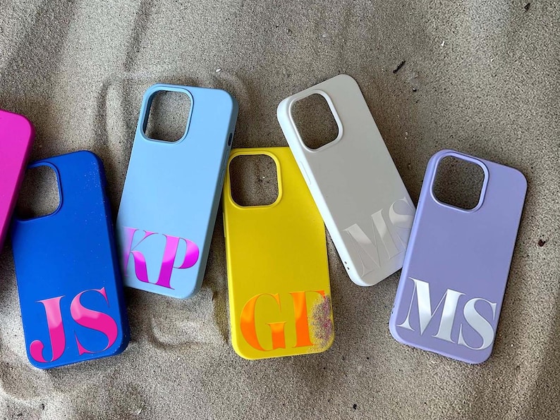 May include: A collection of colorful phone cases in blue, yellow, white, and purple, each with a unique initial design. The cases are arranged on a sandy surface, showcasing a variety of personalized accessories.