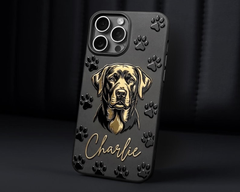 May include: A black smartphone case featuring a raised gold-toned illustration of a dog's face in the center, surrounded by embossed paw prints. The name Charlie is written in a gold cursive script below the dog illustration.