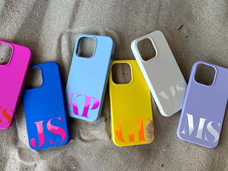 May include: A collection of colorful phone cases in shades of pink, blue, yellow, white, and purple. Each case features a personalized monogram in a contrasting color. The cases are arranged on a sandy surface.