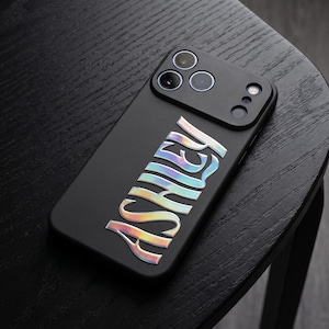 May include: Black phone case with the name "ASHLEY" in iridescent, holographic lettering. The case has a camera module with three lenses and a flash. The phone case is resting on a black wooden surface.
