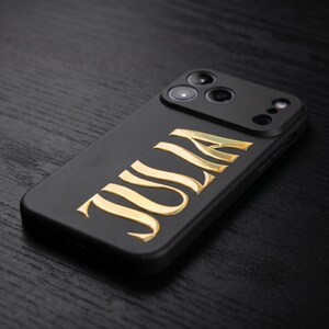 May include: Black phone case with gold-colored lettering spelling out the name "JULIA". The case has a matte finish and a camera module with multiple lenses. The phone case is angled, showing the side profile.
