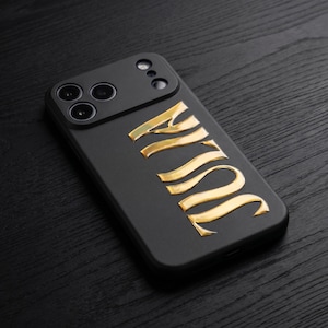 May include: Black phone case with the name "JULIA" in gold lettering. The case has a camera module with three lenses and a flash. The phone case is on a dark wood surface.