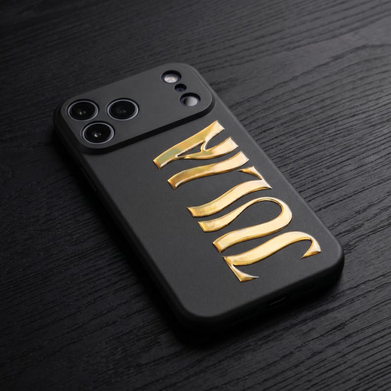 May include: Black phone case with the name "JULIA" in gold lettering. The case has a camera module with three lenses and a flash. The phone case is on a dark wood surface.
