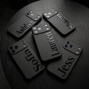 May include: Five matte black smartphone cases arranged in a circular pattern. Each case features a raised, glossy name embossed vertically on the back: Ashley, Sharon, Lauren, Sofia, and Jess. The cases have a cutout for a triple-lens camera system.