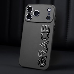 Puede incluir: Una funda de tel&eacute;fono gris oscuro con el nombre "GRACE" en relieve verticalmente. La funda tiene una secci&oacute;n elevada para las lentes de la c&aacute;mara y el flash. El fondo es una superficie oscura y texturizada.