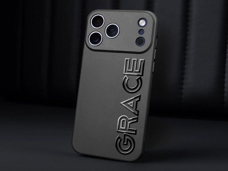 Puede incluir: Una funda de tel&eacute;fono gris oscuro con el nombre "GRACE" en relieve verticalmente. La funda tiene una secci&oacute;n elevada para las lentes de la c&aacute;mara y el flash. El fondo es una superficie oscura y texturizada.