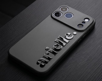 Può includere: Una cover per smartphone nero opaco con il nome arielle. in lettere nere lucide in rilievo. La cover presenta un ritaglio rettangolare per il modulo fotocamera con tre aperture circolari e due aperture più piccole per flash o sensori.
