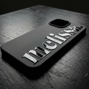 May include: A matte black smartphone case featuring the name Melissa in raised, metallic silver lettering. The case includes a square camera cutout in the upper corner, with the silver text positioned diagonally across the back surface.
