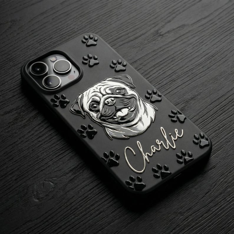 May include: A black smartphone case featuring a raised silver illustration of a pug dog face. The case is decorated with multiple small, raised black paw prints and the name Charlie written in a silver cursive script font on the back surface.
