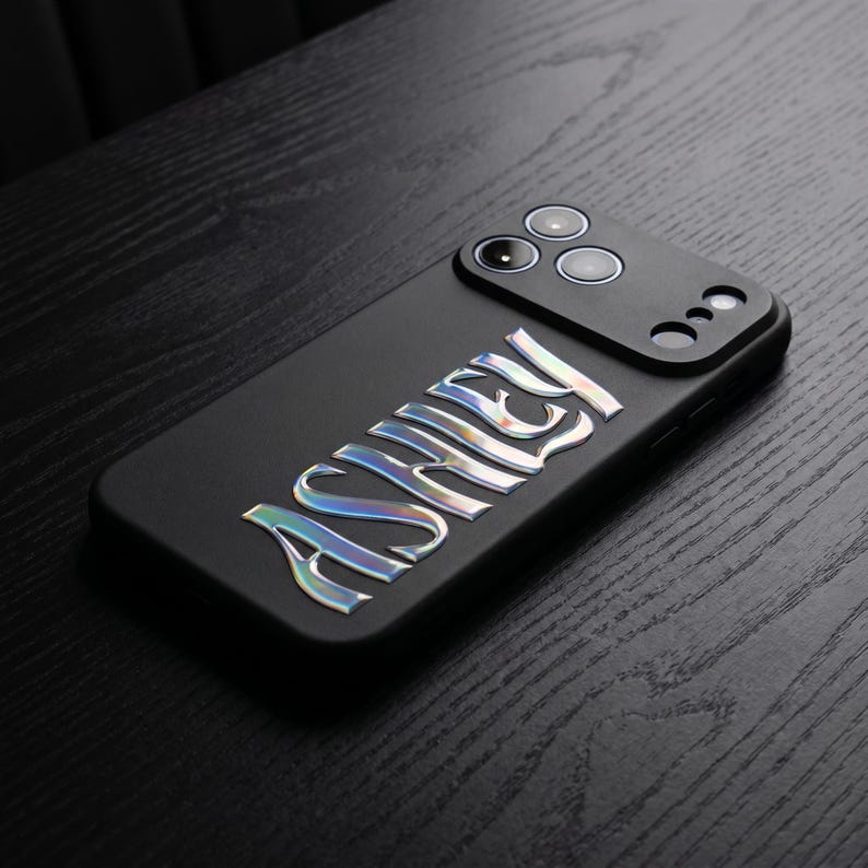 May include: Black phone case with the name "ASHLEY" in iridescent, raised lettering. The case has a camera module with three lenses and a flash. The phone case is on a dark wood surface.
