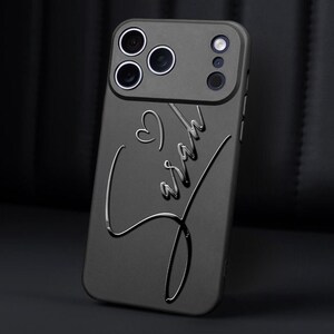 May include: A dark gray phone case with a heart and the name "Sarah" embossed in a cursive font. The case has a camera module with multiple lenses. The overall design is sleek and modern.