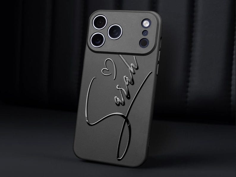 May include: A dark gray phone case with a heart and the name "Sarah" embossed in a cursive font. The case has a camera module with multiple lenses. The overall design is sleek and modern.