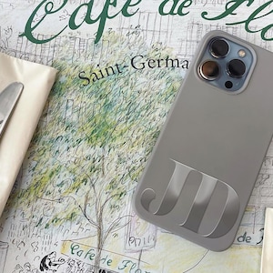 Puede incluir: Una funda gris para tel&eacute;fono con las iniciales "JD" se muestra sobre una superficie similar a un mapa con las palabras "Cafe de Flore" y "Saint-Germa". Un tenedor y un cuchillo descansan sobre una servilleta.