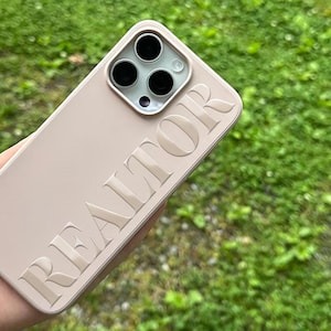 May include: A light pink phone case with the embossed word "REALTOR" in a bold, sans-serif font. The case has a square camera module with three black lenses and a flash. The background is blurred green grass.