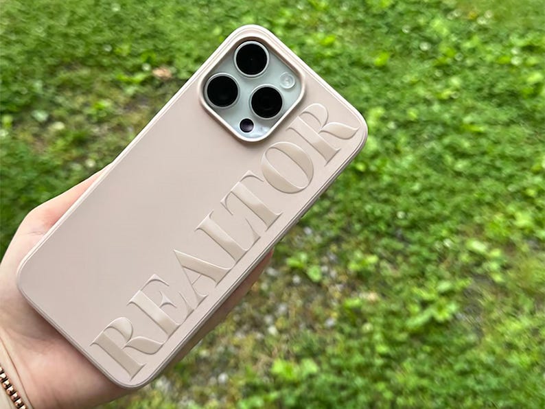 May include: A light pink phone case with the embossed word "REALTOR" in a bold, sans-serif font. The case has a square camera module with three black lenses and a flash. The background is blurred green grass.