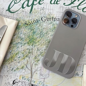 May include: A gray phone case with the initials "JD" in a lighter gray. The phone case is on a table with a napkin, fork, and knife. The background has text that says "Cafe de Flore" and "Saint-Germa".