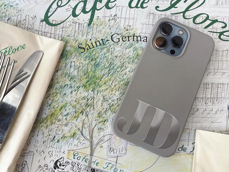 May include: A gray phone case with the initials "JD" in a lighter gray. The phone case is on a table with a napkin, fork, and knife. The background has text that says "Cafe de Flore" and "Saint-Germa".