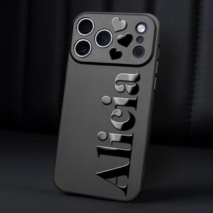 May include: A black phone case with the name "Alicia" embossed in a glossy finish. The case features a camera cutout with heart-shaped accents. The overall design is sleek and modern, suitable for various phone models.