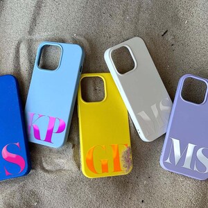 Puede incluir: Una colecci&oacute;n de fundas de tel&eacute;fono personalizadas en varios colores, incluyendo azul, amarillo y lavanda. Cada funda presenta iniciales en un color contrastante, como rosa o naranja. Las fundas est&aacute;n dispuestas sobre una superficie arenosa.
