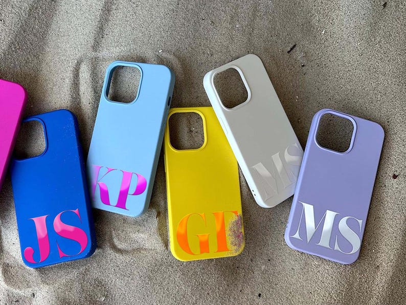 Puede incluir: Una colecci&oacute;n de fundas de tel&eacute;fono personalizadas en varios colores, incluyendo azul, amarillo y lavanda. Cada funda presenta iniciales en un color contrastante, como rosa o naranja. Las fundas est&aacute;n dispuestas sobre una superficie arenosa.