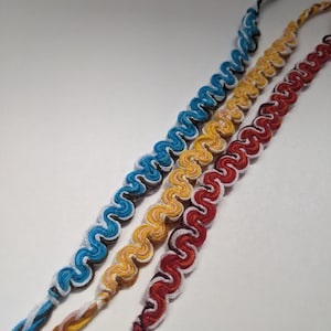 Puede incluir: Tres pulseras de la amistad hechas con hilo de colores. Las pulseras están tejidas en un patrón ondulado y presentan hilo blanco, rojo, amarillo, azul y negro.