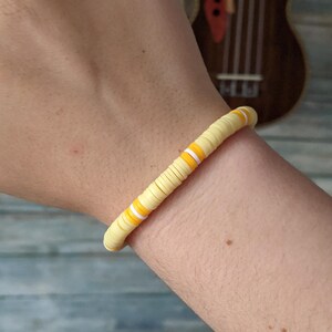 May include: A yellow and white beaded bracelet with orange stripes is worn on a wrist. The bracelet is made of small, flat, circular beads. The background includes a wooden ukulele and a blue wooden surface.