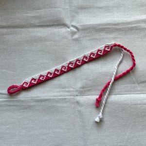 Puede incluir: Pulsera de amistad hecha a mano con un diseño geométrico blanco y rosa. La pulsera está hecha de hilo tejido y tiene un cierre de lazo y lazos trenzados. La pulsera se muestra sobre un fondo de tela blanca.