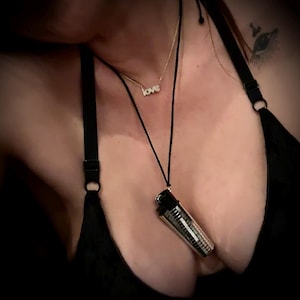 Könnte beinhalten: Eine schwarze Halskette mit einem silbernen Feuerzeug-Anhänger. Eine goldene Halskette mit dem Wort "love" ist ebenfalls sichtbar. Das Bild zeigt eine Person, die ein schwarzes Oberteil trägt.