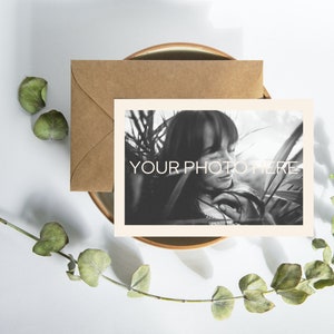 May include: A black and white photograph with the text "YOUR PHOTO HERE" on a cream-colored card, accompanied by a brown envelope. The card and envelope rest on a brown plate, with eucalyptus leaves and stems arranged around them.