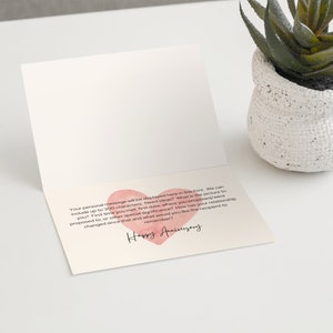 May include: An open greeting card with a pink heart and the words "Happy Anniversary" in cursive. The card is on a white surface next to a small white pot with a green plant.
