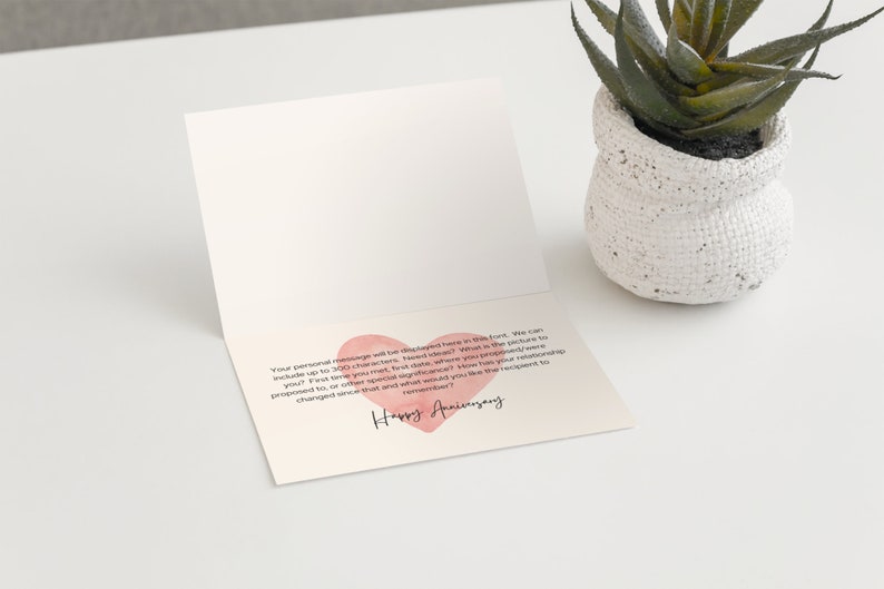 May include: An open greeting card with a pink heart and the words "Happy Anniversary" in cursive. The card is on a white surface next to a small white pot with a green plant.