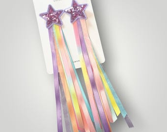May include: Two hair clips on a white card labeled Parker and Kay Collective. Each clip features a purple glitter star with long, hanging satin ribbons in shades of yellow, orange, pink, purple, and light blue, creating a colorful streamer effect.