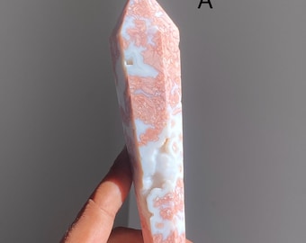 May include: A tall, polished crystal obelisk with a pointed top. The crystal features a pattern of white and pink swirls and patches. The obelisk is held in a hand, and the background is a neutral gray.