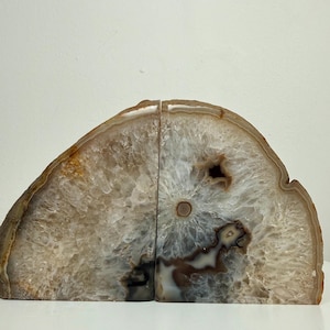 Puede incluir: Dos sujetalibros de ágata. Los sujetalibros están cortados de una piedra natural, mostrando una mezcla de colores marrón, blanco y translúcido. La piedra tiene un patrón en capas y bandeado con una superficie pulida. Los sujetalibros están diseñados para estar de pie.