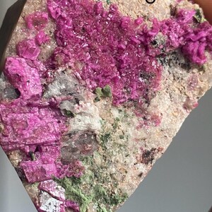 Może przedstawiać: Zbliżenie okazu mineralnego z żywymi, magentowymi formacjami krystalicznymi. Minerał osadzony jest na tle beżowych i zielonych odcieni, z widoczną literą "C" na powierzchni. Okaz trzymany jest w dłoni.