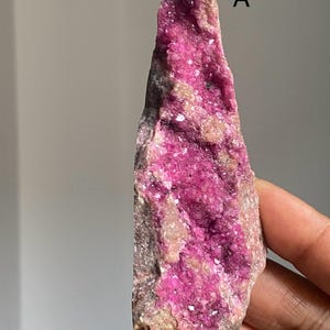 Może przedstawiać: Surowy, naturalny okaz minerału o żywej, intensywnie r&oacute;żowej strukturze krystalicznej. Minerał jest częściowo osadzony w szorstkiej, beżowo-brązowej matrycy. Litera "A" jest widoczna po prawej stronie u g&oacute;ry.