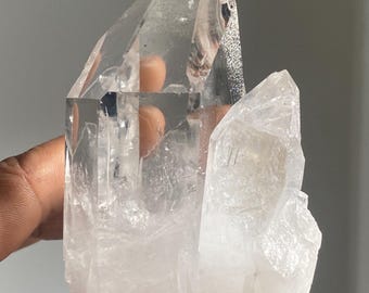 Op de afbeelding: Een heldere kwartskristalgroep met meerdere punten. Het kristal wordt in een hand gehouden, wat de transparante en doorschijnende kwaliteiten laat zien. De kwarts heeft een natuurlijke, ruwe textuur en een kristallijne structuur.