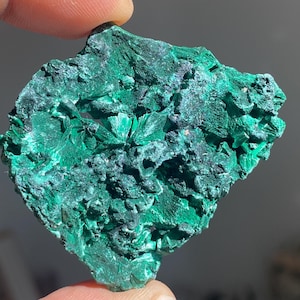 Puede incluir: Una vibrante piedra preciosa de malaquita verde con una superficie rugosa y texturizada. El mineral muestra una estructura cristalina con diferentes tonos de verde, creando un patrón visualmente atractivo. La piedra natural es sostenida por un dedo.