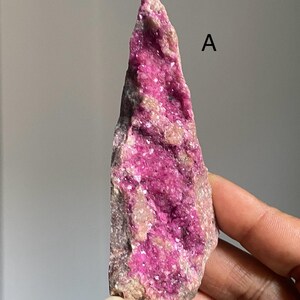 Może przedstawiać: Surowy, naturalny okaz minerału o żywej, intensywnie r&oacute;żowej strukturze krystalicznej. Szorstka, teksturowana powierzchnia prezentuje mieszankę r&oacute;żowych i beżowych odcieni. Widoczna litera "A". Minerał umieszczony na neutralnym tle.