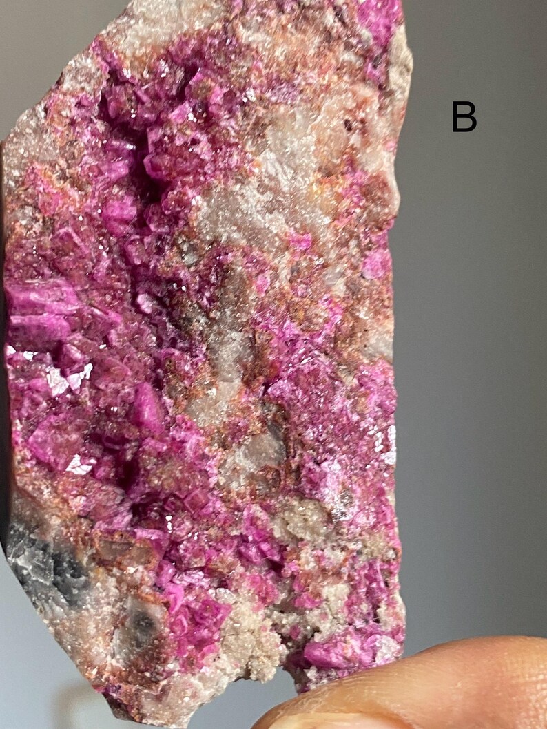 Może przedstawiać: Zbliżenie okazu mineralnego z żywymi, magentowymi formacjami krystalicznymi. Minerał osadzony jest na tle beżowej i szarej skały, z literą "B" widoczną w prawym g&oacute;rnym rogu.