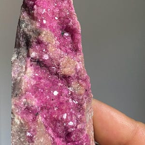Może przedstawiać: Zbliżenie surowego okazu mineralnego o żywej, r&oacute;żowej strukturze krystalicznej. Minerał jest umieszczony na neutralnym tle, prezentując swoją naturalną teksturę i wariacje kolorystyczne. Litera "A" jest widoczna w prawym g&oacute;rnym rogu.