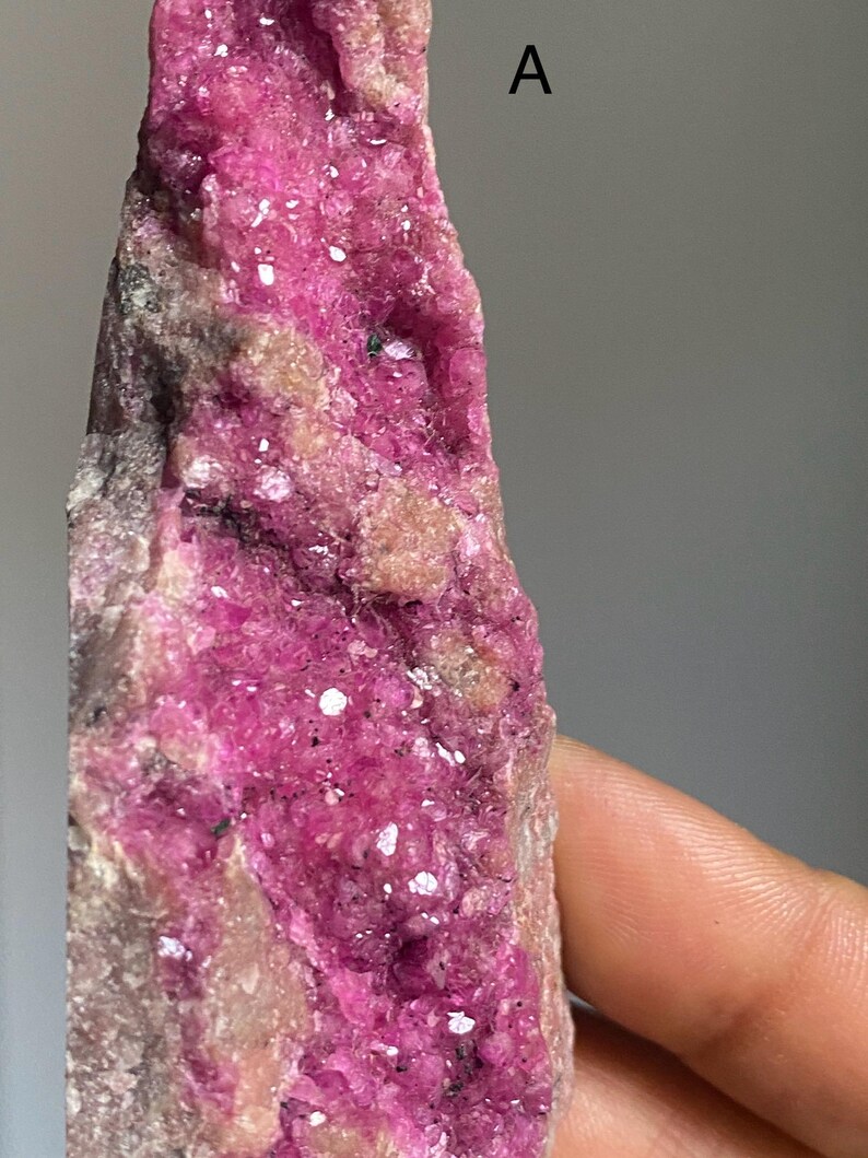 Może przedstawiać: Zbliżenie surowego okazu mineralnego o żywej, r&oacute;żowej strukturze krystalicznej. Minerał jest umieszczony na neutralnym tle, prezentując swoją naturalną teksturę i wariacje kolorystyczne. Litera "A" jest widoczna w prawym g&oacute;rnym rogu.