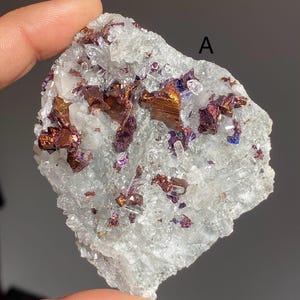 May include: A cluster of iridescent, copper-coloured crystals embedded in a white, translucent quartz matrix. The crystals have a metallic sheen and exhibit a range of colours, including purple, blue, and green. The quartz matrix is rough and textured, with a natural, earthy appearance.