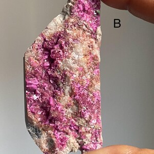 Może przedstawiać: Surowy okaz minerału o nieregularnym kształcie, z żywymi fuksjowymi formacjami krystalicznymi. Matryca kamienia jest jasno-beżowa, z ciemniejszymi szarymi i brązowymi obszarami. Litera "B" jest widoczna po prawej stronie.