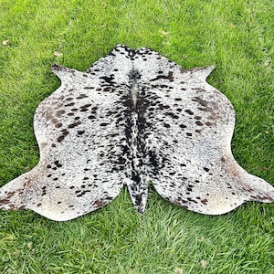以下が含まれることがあります： 白地をベースに黒と茶色の斑点模様が施されたカウハイドラグ。 ラグは不規則な形状で、縁が伸びており、緑の草の上に置かれています。 革は柔らかく、質感のある外観をしています。